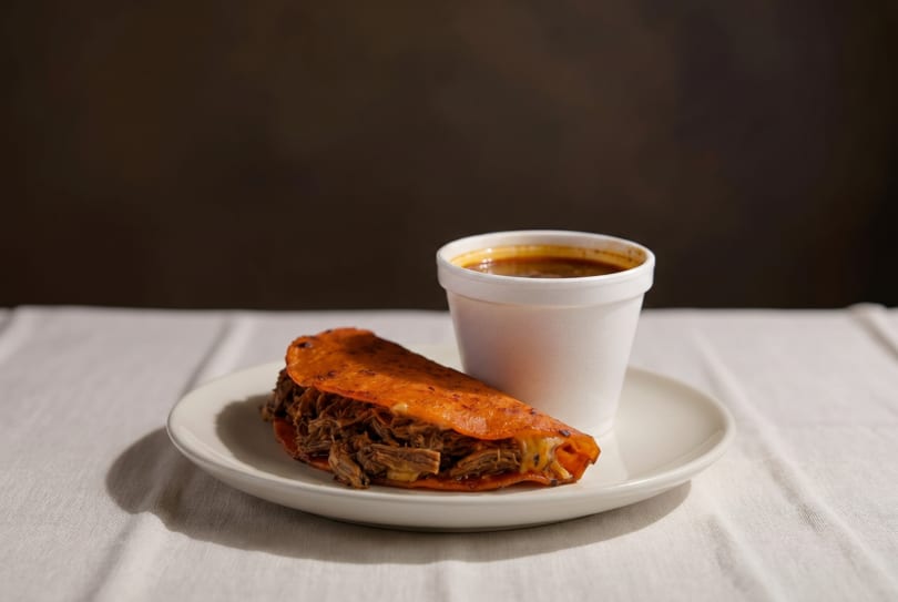 A birria taco with melted cheese and a cup of consommé on a white plate.